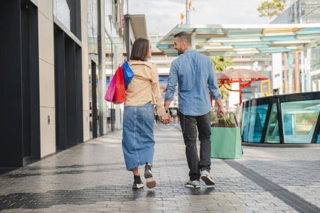 Photo for Back view of young couple shopping on a mall, carrying paper bags in their hands and walking together looking each other and smiling. Husband and wife sightseeing holding hands enjoying on a date - Royalty Free Image