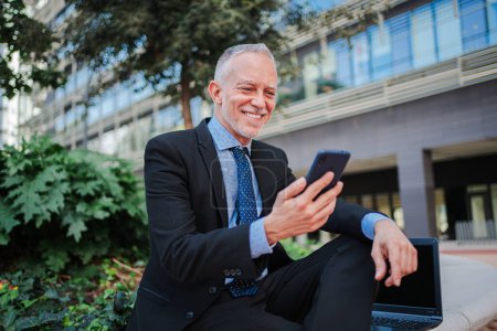 Photo for Smiling mature businessman with suit using a modern smartphone sitting outside relaxing and watching apps. Happy professional executive holding a cell phone and reading a messages at workplace. High - Royalty Free Image