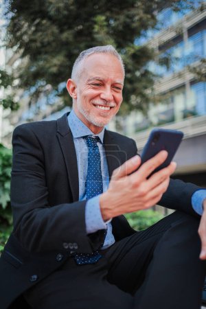 Photo for Vertical. Smiling mature businessman with suit using a smartphone sitting outside relaxing and watching apps. Happy professional executive holding a cell phone and reading a messages at workplace - Royalty Free Image