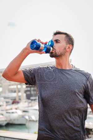 Photo for Vertical. Young handsome sportsman drinking energy water from bottle while resting after run workout. Male person of strong complexion having a break with a supplement and refreshing during training - Royalty Free Image