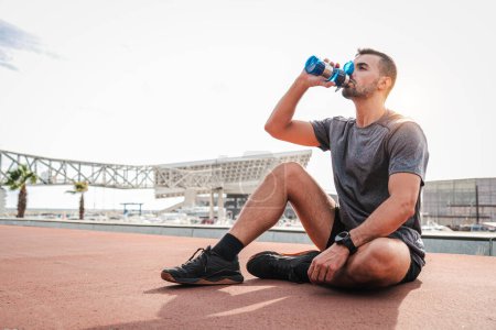 Photo for Weary sports man resting and drinking water after endurance workout sitting on the floor. Exhauted male recovering and having a routine break taking a protein supplement at cardio wellness training - Royalty Free Image