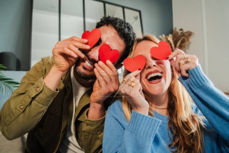 Téléchargez les photos : Jeune couple souriant couvrant leurs yeux avec des papiers en forme de coeur rouge, s'amusant à la Saint-Valentin. Petit ami et petite amie célébrant leur relation amoureuse ensemble à un rendez-vous romantique à la maison - en image libre de droit