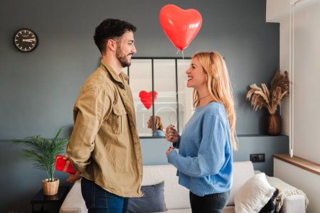 Photo for Boyfriend giving a valentines day suprise present to his girlfriend, she is holding a heart shape inflated balloon. Young couple celebrating a valentine holiday with romantic gifts. Romance and love - Royalty Free Image