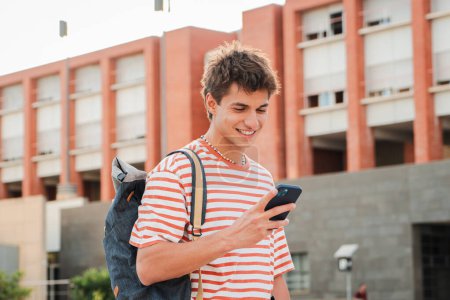 Foto de Estudiante masculino divirtiéndose con un teléfono celular publicando en las redes sociales en el campus universitario. Hombre adolescente real sonriendo con un teléfono inteligente. Joven sosteniendo un teléfono móvil. Adolescente escolar leyendo un teléfono - Imagen libre de derechos