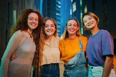 Téléchargez les photos : Portrait d'un groupe de jeunes femmes souriantes regardant ensemble la caméra la nuit. Quatre amies dehors avec une expression heureuse. Fières dames féminines regardant devant le rassemblement social - en image libre de droit