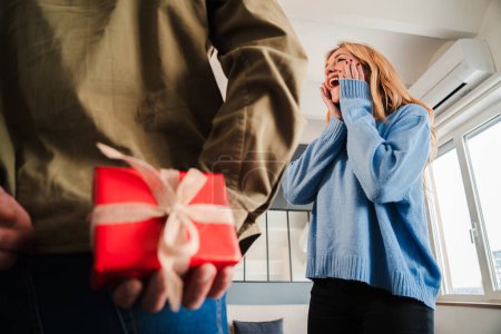 Photo for Rear view of a man hiding a valentine surprise gift to his girlfriend. Couple celebrating a valentines day holiday. Surprised wife watching a birthday present from her husband. Man giving a giftbox - Royalty Free Image