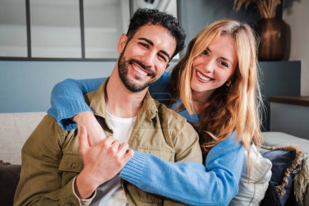 Photo for Close up portrait of a young caucasian married couple smiling together and looking at camera sitting at home sofa. Affectionate wife embracing her handsome husband at living room couch. Relationship - Royalty Free Image