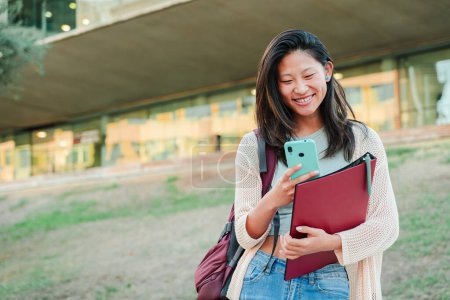 Téléchargez les photos : Une écolière chinoise souriant à l'aide d'un téléphone portable pour partager des photos sur l'application de médias sociaux du campus universitaire tenant un sac à dos et un carnet. Étudiante adolescente heureuse avec un téléphone intelligent. Haut - en image libre de droit