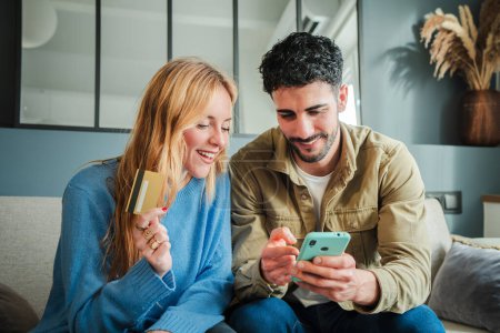 Photo for A couple is sharing a moment of leisure on the couch at home, looking at a cell phone with smiles. They seem happy and relaxed during this fun event, using technology and a credit card to buy online - Royalty Free Image