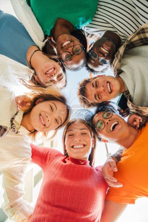 Foto de Vista vertical de bajo ángulo de un grupo de amigos multirraciales parados en un círculo, sonriendo y abrazándose juntos. Jóvenes adolescentes riendo y mirando a la cámara. Equipo de personas en una reunión de coaching - Imagen libre de derechos