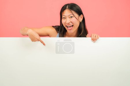 Photo for Young asian woman pointing a blank empty board against red backdrop. Advice copy space mockup. Chinese teenage girl showing a down billboard copyspace at pink color background wall. Female presenting - Royalty Free Image