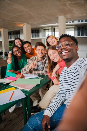 Foto de Jóvenes estudiantes felices tomando un retrato vertical de selfie juntos en la biblioteca universitaria. Chico afroamericano tomando una foto con sus compañeros sonrientes en una reunión de la escuela secundaria. Amigos en la academia - Imagen libre de derechos