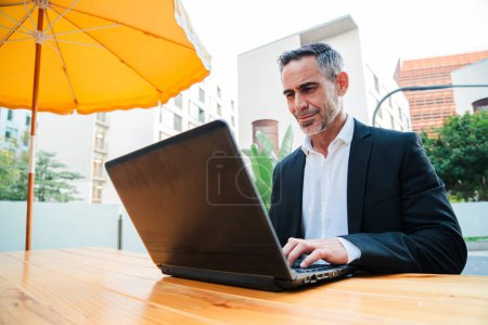 Téléchargez les photos : Homme d'affaires mature travaillant avec un ordinateur portable à l'extérieur, assis au café. Beau cadre masculin utilisant un ordinateur sans fil pour opérer sur le marché boursier. Dactylographie réussie des cols blancs. Haut - en image libre de droit
