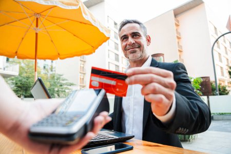 Photo for Happy mature business man paying bill using a contactless credit card in a restaurant. Handsome mid adult male customer smiling holding a creditcard and giving a payment transaction to the cashier - Royalty Free Image