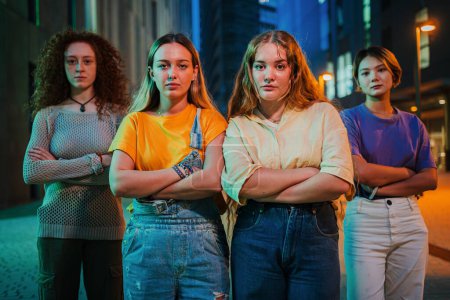 Portrait of a group of serious young women looking at camera together at night time. Proud feminine girls staring front at social gathering. Four real female friends standing outside with pensive