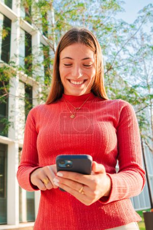 Photo for Vertical portrait of smiling real young woman using mobile phone app , texting, playing game, shopping online, ordering delivery. Happy blonde teen girl sharing social media holding smartphone. High - Royalty Free Image
