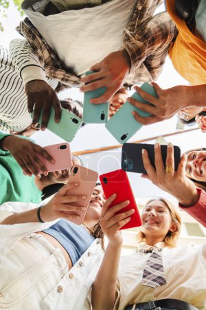 Photo for Vertical low angle view of a big group of smiling multiracial teenagers addicted to smart phones, watching funny videos, shopping online, enjoying outdoors. Young real people using their cellphones - Royalty Free Image