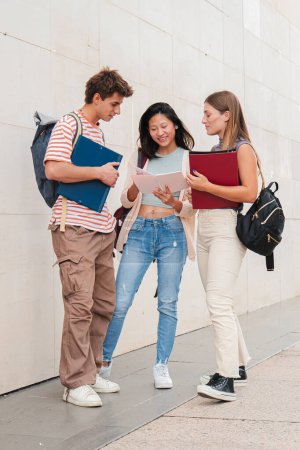 Photo for Vertical. Group of classmates talking about the class, standing at university campus. Three real students having a friendly conversation about the high school homework. Young academy people speaking - Royalty Free Image