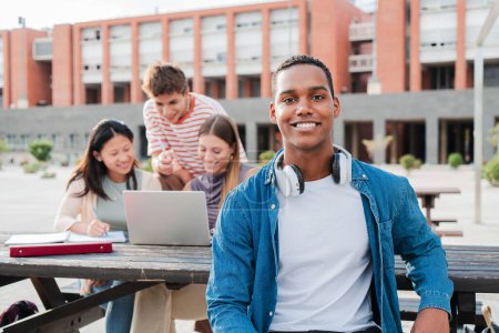 Téléchargez les photos : Jeune étudiant afro-américain, souriant et regardant la caméra assise avec ses camarades de classe sur le campus universitaire. écolier fixer devant derrière son multiracial amis alors qu'ils utilisent un ordinateur portable - en image libre de droit