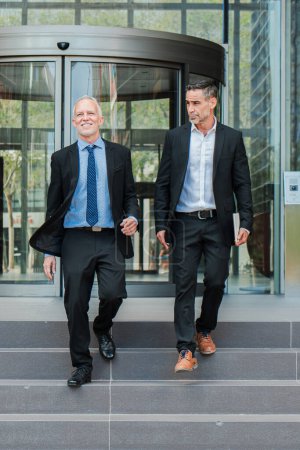Photo for Vertical. Confident business men walking down the staircase, talking about the corporate finance. Office colleagues on a stairway having a conversation. Middle aged executive males discurssing. High - Royalty Free Image
