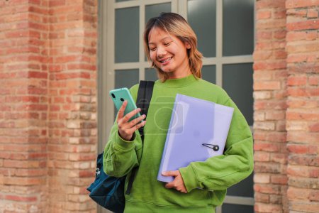 Téléchargez les photos : Jeune fille asiatique lycéenne souriant en utilisant son téléphone portable. Chinoise adolescente naviguant sur Internet avec une application smartphone sur le campus universitaire. Thaï camarade de classe femme regarder un téléphone mobile - en image libre de droit