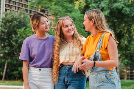 Photo for Three teenage gossip girls talking together outside. Group of multiracial female friends chatting and having communication. Young adult student women smiling and walking together on a social meeting - Royalty Free Image