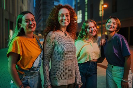 Porträt einer Gruppe junger erwachsener Frauen, die nachts zusammen stehen. Vier Mädchen im Teenageralter lächeln zusammen und blicken in die Kamera eines gesellschaftlichen Treffens. Echte Damen lachen und starren um Mitternacht nach vorne