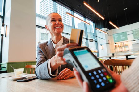 Photo for Happy mid adult woman paying the bill with a contactless credit card in a restaurant. Female smiling holding a creditcard and giving a payment transaction to the cashier dataphone on a coffee shop - Royalty Free Image