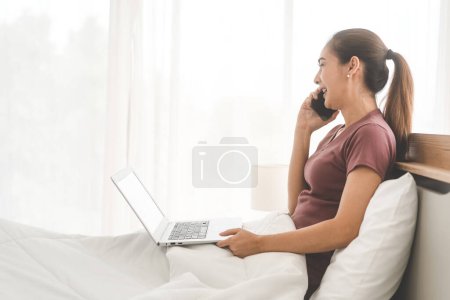 Lifestyle-Routine mit Internet-Technologie-Konzept. Junge erwachsene asiatische Frau mit Laptop und sprechen mit Smartphone auf dem Bett für telemedizinische psychische Gesundheit. Menschen ruhen sich im Schlafzimmer aus.