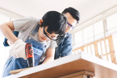 Foto de Southeast asian family father and son diy activity together at home concept. Boy using drill for learn skill of carpenter with parent at workshop. - Imagen libre de derechos