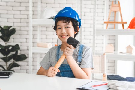 Foto de Southeast asian child self education for home school. Boy wearing safety helmet and holding hammer for engineer dream future. - Imagen libre de derechos
