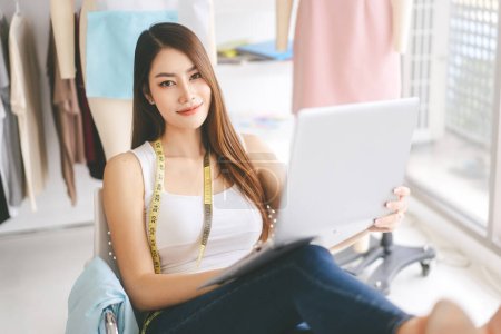 Photo for People tailor working in fashion design concept. Portrait of beautiful young adult asian woman designer sitting using laptop and eye looking camera in workshop studio background. - Royalty Free Image