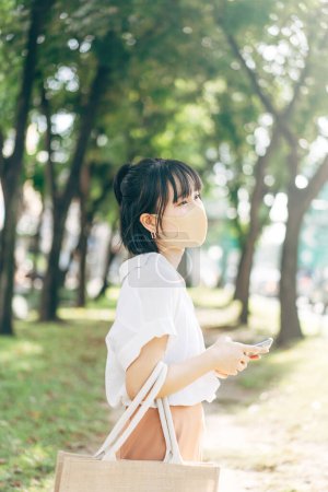 Téléchargez les photos : Young adult business asian working woman wear face mask for protect virus covid19. Urban people lifestyle after got vaccine on day concept. Public park with tree and street background. - en image libre de droit