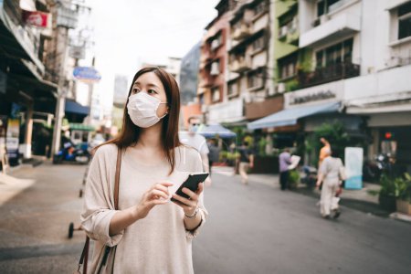 Téléchargez les photos : Casual business young asian single woman wear mask for new normal lifestyle protect corona virus or covid19. Using smartphone on sidewalk street outdoor area on day. Bangkok, Thailand. - en image libre de droit