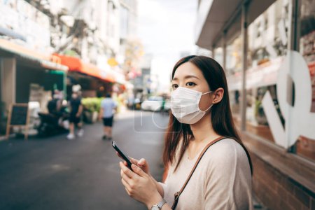 Téléchargez les photos : Casual business young asian single woman wear face mask for new normal lifestyle protect corona virus or covid19. Using smartphone on sidewalk street outdoor area on day. Bangkok, Thailand. - en image libre de droit