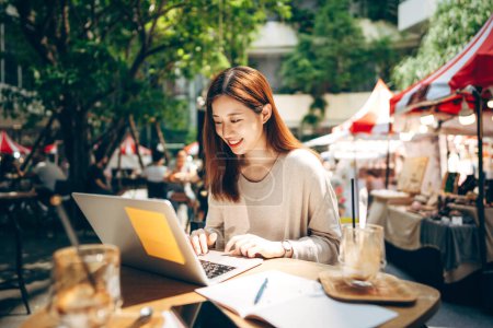 Téléchargez les photos : Entreprise pigiste adulte asiatique femme utilisant un ordinateur portable pour le travail au café trottoir. Style de vie urbain avec la technologie moderne sur le jour. - en image libre de droit