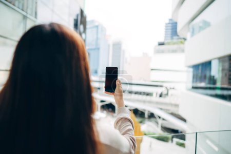 Foto de Shoulder view of woman holding mobile phone empty screen with blur city background on day. - Imagen libre de derechos