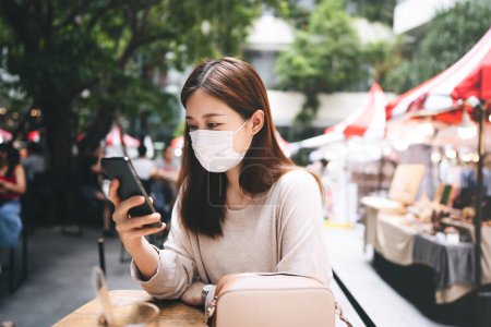 Photo for Young adult asian female with protective face mask for virus corona or covid 19. Using mobile phone for online application. Urban lifestyle people at cafeteria on day - Royalty Free Image