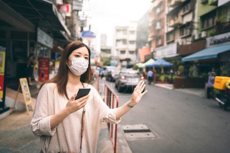 Foto de Negocios casuales joven asiático mujer soltera usar mascarilla para el nuevo estilo de vida normal proteger el virus de la corona o COVID-19. Uso de la aplicación de teléfono inteligente para llamar taxi en el día. Bangkok, Tailandia. - Imagen libre de derechos