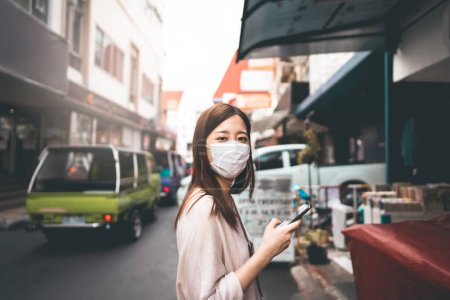 Foto de Negocios casuales joven asiático mujer soltera usar mascarilla para el nuevo estilo de vida normal proteger el virus de la corona o COVID-19. El uso de teléfono inteligente en la acera calle zona al aire libre en el día. Bangkok, Tailandia. - Imagen libre de derechos