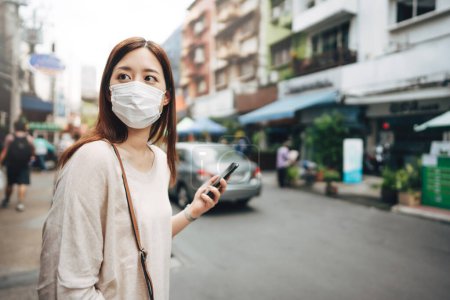Foto de Casual business young asian single woman wear face mask for new normal lifestyle protect corona virus or COVID-19. Using smartphone on sidewalk street outdoor area on day. Bangkok, Thailand. - Imagen libre de derechos