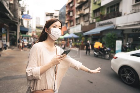Téléchargez les photos : Casual business young asian single woman wear face mask for new normal lifestyle protect corona virus or COVID-19. Using smartphone application for call taxi on day. Bangkok, Thailand. - en image libre de droit