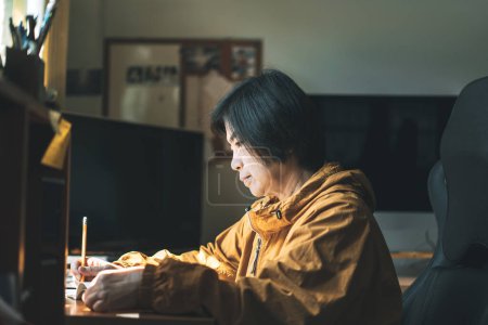Téléchargez les photos : Alone asian elder woman work writing in the morning at workplace. Senior self care with home hobby concept. Warm light from window. - en image libre de droit