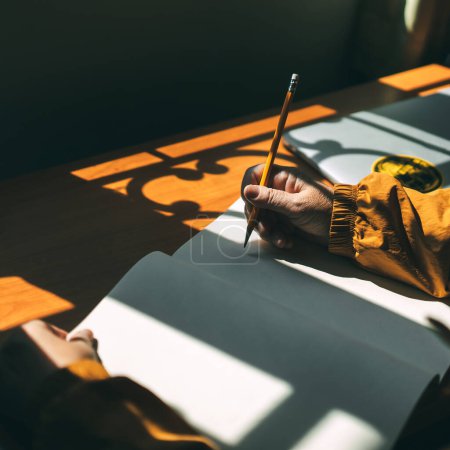 Photo for Elder hand hold pencil writing with sun light and shadow on wood table. Warm and yellow mood. - Royalty Free Image