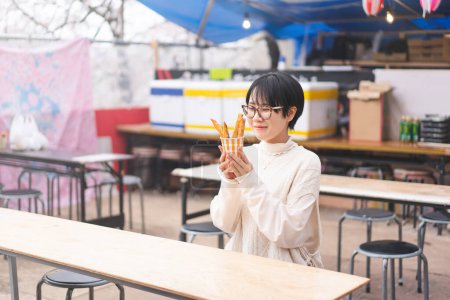 Foto de Feliz sonrisa joven adulto asiático turista mujer sentarse y comer japonés estilo calle comida. Japón viaje temporada con sakura cerezo flor árbol primavera festival. - Imagen libre de derechos