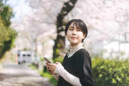 Photo for Young adult asian woman using smartphone. Typing for online dating app or social media. Japanese people lifestyles at park outdoor on spring season. Background with copy space. - Royalty Free Image