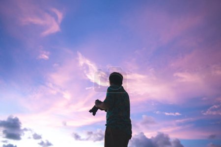 Téléchargez les photos : Silhouette vue arrière du photographe asiatique de voyage adulte homme avec appareil photo sous le beau ciel spectaculaire coucher de soleil. Les gens se reposent et explorent la thérapie avec l'environnement naturel. - en image libre de droit
