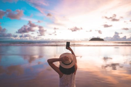 Photo for Rear view of young adult tourist asian woman walking relax on beach sand using smartphone for take photo. Outdoor domestic travel at southeast asia andaman ocean. Phuket, Thailand. - Royalty Free Image