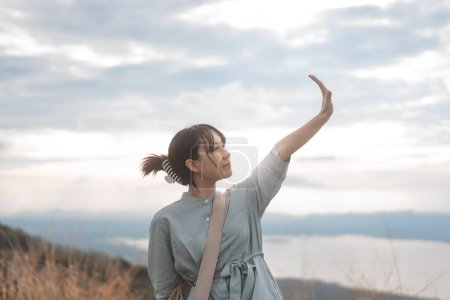 Téléchargez les photos : Jeune adulte asiatique voyageur femme vue de côté levant la main vers le ciel. Debout à l'extérieur se détendre avec la nature sentir la liberté et l'âme. - en image libre de droit