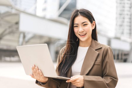 Téléchargez les photos : Entreprise indépendante jeune femme asiatique adulte utilisant un ordinateur portable pour le travail. Beau sourire visage cheveux longs. Assis à l'extérieur le jour. Urbain style de vie urbain. Fille regardant caméra. - en image libre de droit